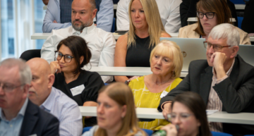 A photograph of an audience seated in tiered rows at a conference or lecture setting. Several adults are visible across multiple rows, facing forward and listening to a speaker who is not shown. In the centre row, a person in a sleeveless dark top sits next to a person in a white shirt, while another attendee to the right is working on a laptop with a takeaway coffee cup nearby. In the row in front, a person in a bright yellow top sits next to someone in a dark blazer. Other attendees are visible in the foreground and background, some slightly out of focus. The room features white desks and blue seating, with bright, even lighting typical of a conference or seminar environment.