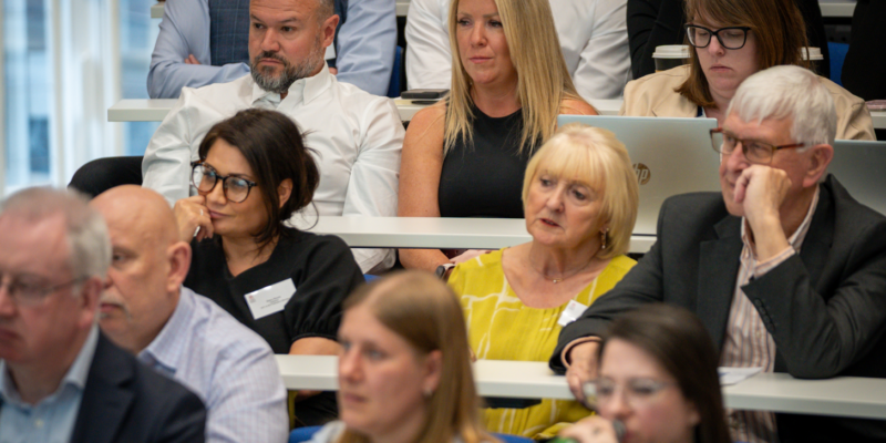A photograph of an audience seated in tiered rows at a conference or lecture setting. Several adults are visible across multiple rows, facing forward and listening to a speaker who is not shown. In the centre row, a person in a sleeveless dark top sits next to a person in a white shirt, while another attendee to the right is working on a laptop with a takeaway coffee cup nearby. In the row in front, a person in a bright yellow top sits next to someone in a dark blazer. Other attendees are visible in the foreground and background, some slightly out of focus. The room features white desks and blue seating, with bright, even lighting typical of a conference or seminar environment.
