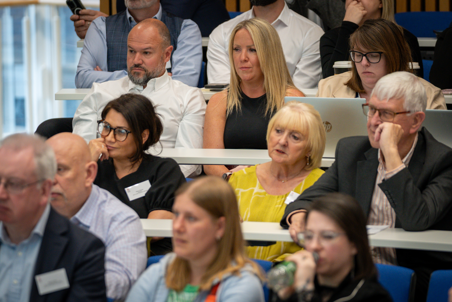 A photograph of an audience seated in tiered rows at a conference or lecture setting. Several adults are visible across multiple rows, facing forward and listening to a speaker who is not shown. In the centre row, a person in a sleeveless dark top sits next to a person in a white shirt, while another attendee to the right is working on a laptop with a takeaway coffee cup nearby. In the row in front, a person in a bright yellow top sits next to someone in a dark blazer. Other attendees are visible in the foreground and background, some slightly out of focus. The room features white desks and blue seating, with bright, even lighting typical of a conference or seminar environment.